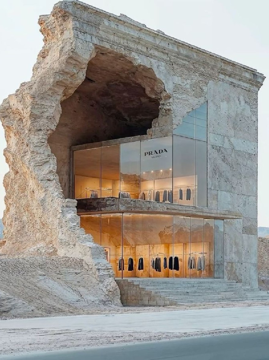 Русский промпт: A rendering of a prada store in the desert, featuring a large, eroded rock formation with a large opening at the entrance. The store is made of light-colored stone and has a glass facade with the Prada logo prominently displayed. Inside the store, clothes are hung on hangers, and the store is surrounded by barren desert landscape with mountains in the background. The sky is clear and the lighting is soft, casting a warm glow on the clothing. The image has a minimalist design with clean lines and a minimalist aesthetic. | Original prompt: A rendering of a prada store in the desert, featuring a large, eroded rock formation with a large opening at the entrance. The store is made of light-colored stone and has a glass facade with the Prada logo prominently displayed. Inside the store, clothes are hung on hangers, and the store is surrounded by barren desert landscape with mountains in the background. The sky is clear and the lighting is soft, casting a warm glow on the clothing. The image has a minimalist design with clean lines and a minimalist aesthetic.
