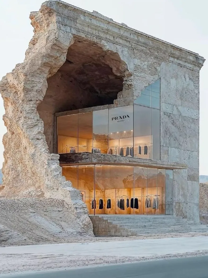 Русский промпт: A rendering of a prada store in the desert, featuring a large, eroded rock formation with a large opening at the entrance. The store is made of light-colored stone and has a glass facade with the Prada logo prominently displayed. Inside the store, clothes are hung on hangers, and the store is surrounded by barren desert landscape with mountains in the background. The sky is clear and the lighting is soft, casting a warm glow on the clothing. The image has a minimalist design with clean lines and a minimalist aesthetic. | Original prompt: A rendering of a prada store in the desert, featuring a large, eroded rock formation with a large opening at the entrance. The store is made of light-colored stone and has a glass facade with the Prada logo prominently displayed. Inside the store, clothes are hung on hangers, and the store is surrounded by barren desert landscape with mountains in the background. The sky is clear and the lighting is soft, casting a warm glow on the clothing. The image has a minimalist design with clean lines and a minimalist aesthetic.
