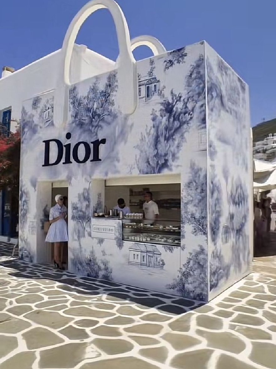 Русский промпт: A rendering of a Dior store in Santorini, Greece, featuring a white building with blue and white toile-patterned walls and a large Dior bag-shaped window display. The store is situated on a cobblestone street, with a bright blue sky in the background. Two people are standing in front of the store, one wearing a white dress and the other wearing a blue dress, both looking at the display. There is a watermark in the bottom right corner of the image. | Original prompt: A rendering of a Dior store in Santorini, Greece, featuring a white building with blue and white toile-patterned walls and a large Dior bag-shaped window display. The store is situated on a cobblestone street, with a bright blue sky in the background. Two people are standing in front of the store, one wearing a white dress and the other wearing a blue dress, both looking at the display. There is a watermark in the bottom right corner of the image.