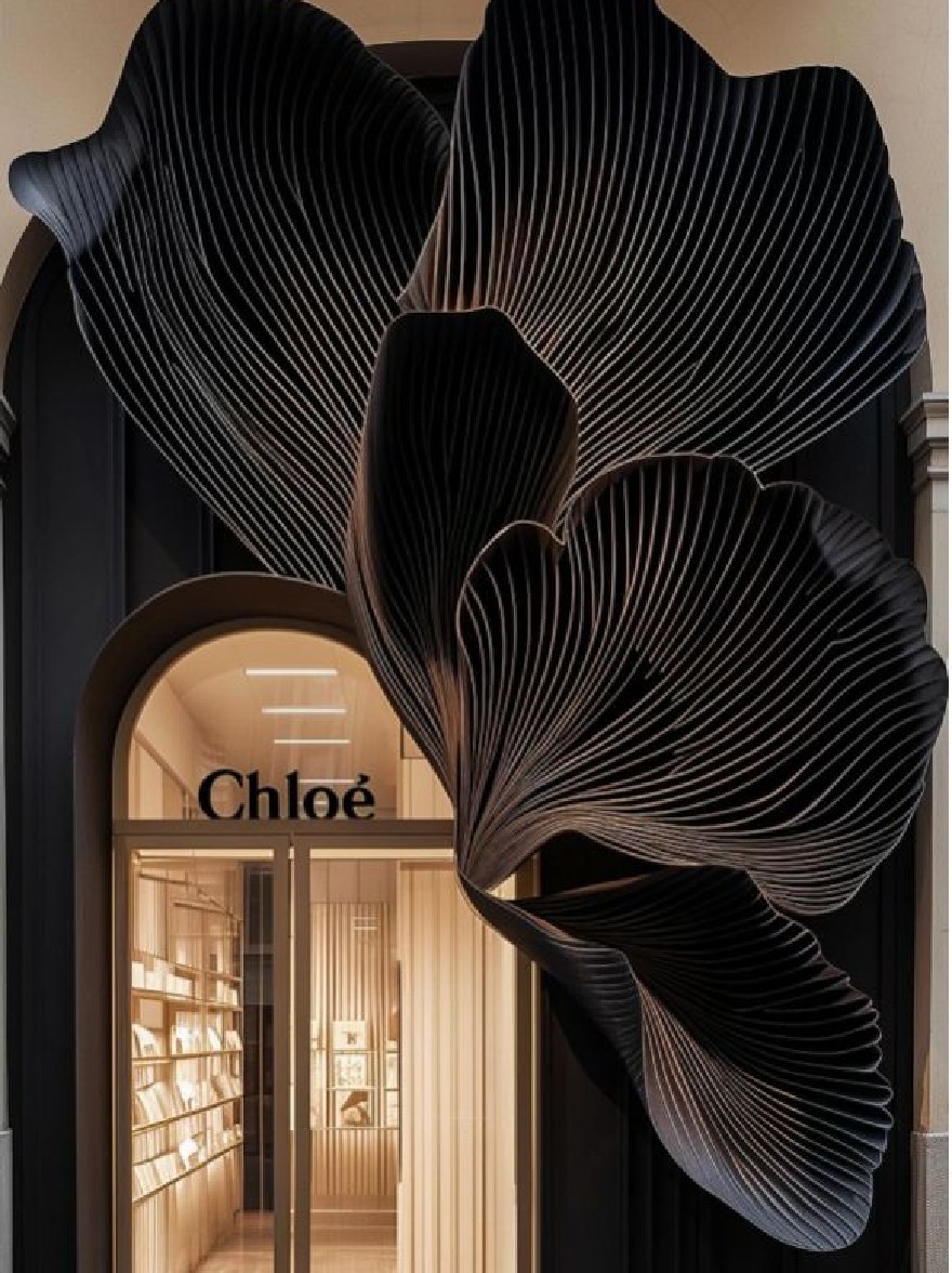 Русский промпт: The image shows the entrance of a Chloé store in Paris, France, with a large, black, leaf-like sculpture mounted on the wall above the entrance. The sculpture is intricately designed with thin, wavy lines that create a textured surface. The entrance is framed by a large glass door that leads into the store, with the word "Chloé" written in white cursive letters above it. The lighting is dim, creating a warm, inviting atmosphere. The walls are painted in a neutral color, and the floor is made of light-colored wood. The overall aesthetic of the store is modern and sophisticated. | Original prompt: The image shows the entrance of a Chloé store in Paris, France, with a large, black, leaf-like sculpture mounted on the wall above the entrance. The sculpture is intricately designed with thin, wavy lines that create a textured surface. The entrance is framed by a large glass door that leads into the store, with the word "Chloé" written in white cursive letters above it. The lighting is dim, creating a warm, inviting atmosphere. The walls are painted in a neutral color, and the floor is made of light-colored wood. The overall aesthetic of the store is modern and sophisticated.