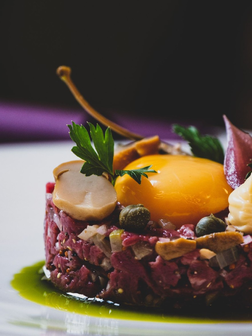 Русский промпт: Фотография тарелки из сырого говяжьего тартара с жареным яйцом сверху, украшенная нарезанными грибами, солеными огурцами и веточкой петрушки. Тартарь подается на белой тарелке с зеленым соусом, сбрызующим его. Фон размыт, но это, кажется, находится в помещении с фиолетовой тканью, наброшенной над краем. Освещение мягкое и естественное, выделяя тартар и ингредиенты. Общая эстетика элегантная и сложная. | Original prompt: A photograph of a plate of raw beef tartare with a fried egg on top, garnished with sliced mushrooms, pickles, and a sprig of parsley. The tartare is served on a white plate with a green sauce drizzled over it. The background is blurred, but it appears to be an indoor setting with a purple cloth draped over the edge. The lighting is soft and natural, highlighting the tartare and the ingredients. The overall aesthetic is elegant and sophisticated.