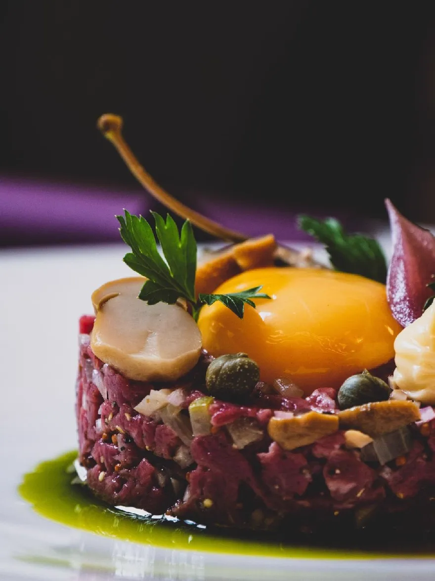 Русский промпт: Фотография тарелки из сырого говяжьего тартара с жареным яйцом сверху, украшенная нарезанными грибами, солеными огурцами и веточкой петрушки. Тартарь подается на белой тарелке с зеленым соусом, сбрызующим его. Фон размыт, но это, кажется, находится в помещении с фиолетовой тканью, наброшенной над краем. Освещение мягкое и естественное, выделяя тартар и ингредиенты. Общая эстетика элегантная и сложная. | Original prompt: A photograph of a plate of raw beef tartare with a fried egg on top, garnished with sliced mushrooms, pickles, and a sprig of parsley. The tartare is served on a white plate with a green sauce drizzled over it. The background is blurred, but it appears to be an indoor setting with a purple cloth draped over the edge. The lighting is soft and natural, highlighting the tartare and the ingredients. The overall aesthetic is elegant and sophisticated.