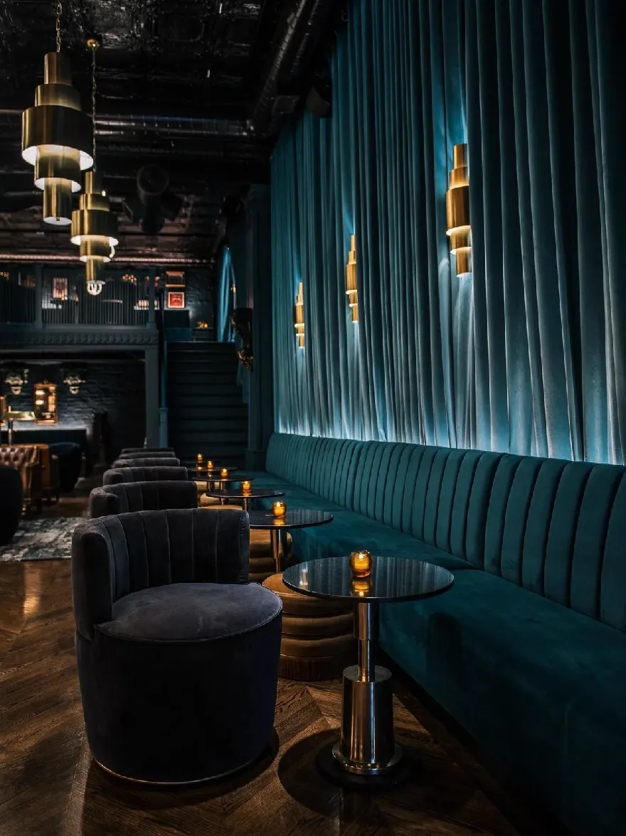 Русский промпт: Тусклонно освещенная, роскошная гостиная с темно-синим бархатным диваном и круглыми, черными бархатными стульями, расположенными в полукруге вокруг стеклянного стола в центре комнаты. Стены украшены золотыми лампами, создавая теплую и привлекательную атмосферу. В комнате есть деревенское промышленное ощущение с открытыми кирпичными стенами и темными деревянными полами. Освещение создает драматический эффект, с синей бархатной драпировкой, обеспечивающей мягкую, бархатистую текстуру. Общая атмосфера утончена и привлекательна, идеально подходит для расслабляющего вечера. | Original prompt: A dimly lit, luxurious lounge with dark blue velvet sofas and round, black velvet chairs arranged in a semi-circle around a glass table in the center of the room. The walls are adorned with gold-plated lamps, creating a warm and inviting atmosphere. The room has a rustic, industrial feel with exposed brick walls and dark wood floors. The lighting creates a dramatic effect, with the blue velvet drapery providing a soft, velvety texture. The overall ambiance is sophisticated and inviting, perfect for a relaxing evening.