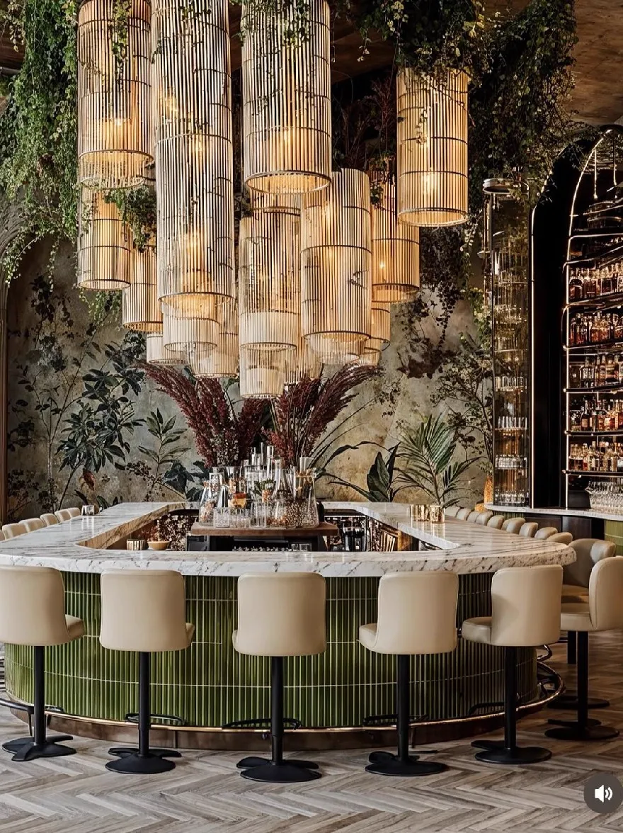 Русский промпт: На изображении показан роскошный бар с большой круглой мраморной столешницей в центре, окруженном бежевыми кожаными барными классами. Стременная столешница украшена зеленым бамбуковым ковриком и имеет современный минималистский дизайн. Над столешницей большая люстра, сделанная из бамбуковых палочек, висит с потолка, с пышной зеленью, каскадной по стенам. На правой стороне изображения высокий цилиндрический витрин, заполненный бутылками ликера, стоит за стержней, добавляя оттенки изысканности. Освещение тепло и привлекательно, создавая уютную атмосферу. Пол изготовлен из плитки елочки, добавляя текстуру и глубину в пространство. | Original prompt: The image shows a luxurious bar with a large, round, marble countertop in the center, surrounded by beige leather barstools. The countertop is decorated with a green bamboo mat and has a modern, minimalist design. Above the countertop, a large chandelier made of bamboo sticks hangs from the ceiling, with lush greenery cascading down the walls. On the right side of the image, a tall, cylindrical display case filled with bottles of liquor stands behind the bar, adding a touch of sophistication. The lighting is warm and inviting, creating a cozy atmosphere. The floor is made of herringbone tiles, adding texture and depth to the space.