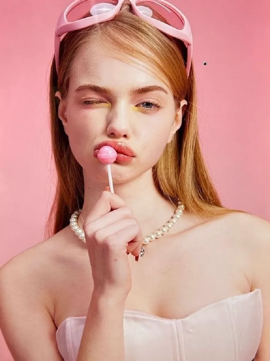 Русский промпт: Photo of a young woman with long, straight, light brown hair, wearing a white strapless dress and a pearl necklace. She is holding a pink lollipop in her right hand, with her left hand resting on her chin. Her eyes are painted with bright yellow eyeshadow, and she has a neutral expression. The background is a solid pink color. The lighting is soft and even, highlighting her delicate features. The overall aesthetic is minimalistic and modern. | Original prompt: Photo of a young woman with long, straight, light brown hair, wearing a white strapless dress and a pearl necklace. She is holding a pink lollipop in her right hand, with her left hand resting on her chin. Her eyes are painted with bright yellow eyeshadow, and she has a neutral expression. The background is a solid pink color. The lighting is soft and even, highlighting her delicate features. The overall aesthetic is minimalistic and modern.