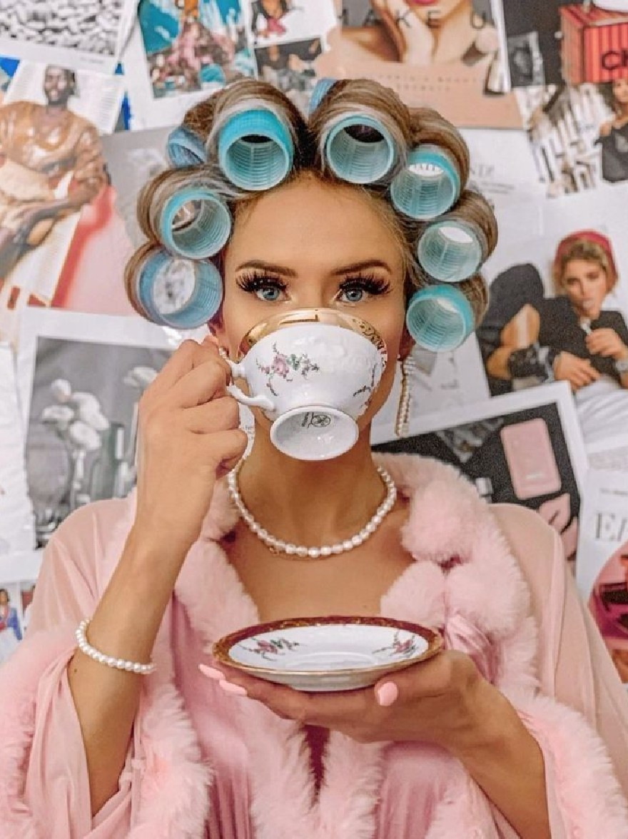 Русский промпт: Photo of a young woman with curlers on her head, holding a teacup and saucer in front of her face. She is wearing a pink fur stole draped over her shoulders and a pearl necklace. The background is a collage of various photographs and magazines. The woman has a neutral expression and is looking directly at the camera. The lighting is soft and natural, highlighting her features. The overall aesthetic is vintage and whimsical. | Original prompt: Photo of a young woman with curlers on her head, holding a teacup and saucer in front of her face. She is wearing a pink fur stole draped over her shoulders and a pearl necklace. The background is a collage of various photographs and magazines. The woman has a neutral expression and is looking directly at the camera. The lighting is soft and natural, highlighting her features. The overall aesthetic is vintage and whimsical.