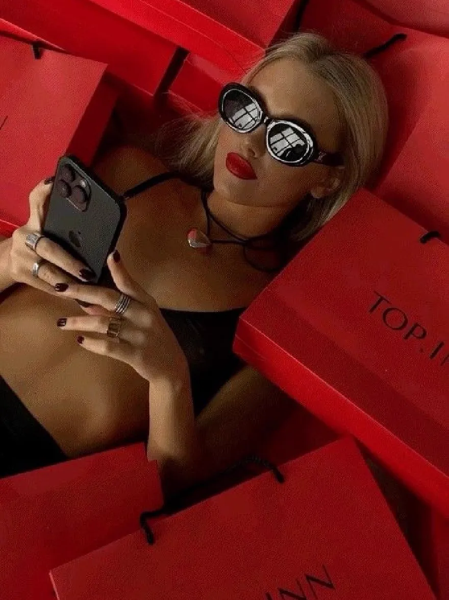 Русский промпт: Photo of a woman lying in a pile of red shopping bags, wearing a black crop top and holding a black iPhone in her hands. She has blonde hair, red lipstick, and black sunglasses. The bags have the words "TOP.INN" written on them in white. She is wearing multiple rings on her fingers. The background is a bright red color. The lighting is dramatic, highlighting her face and the bags. The image has a high-quality, professional look. | Original prompt: Photo of a woman lying in a pile of red shopping bags, wearing a black crop top and holding a black iPhone in her hands. She has blonde hair, red lipstick, and black sunglasses. The bags have the words "TOP.INN" written on them in white. She is wearing multiple rings on her fingers. The background is a bright red color. The lighting is dramatic, highlighting her face and the bags. The image has a high-quality, professional look.