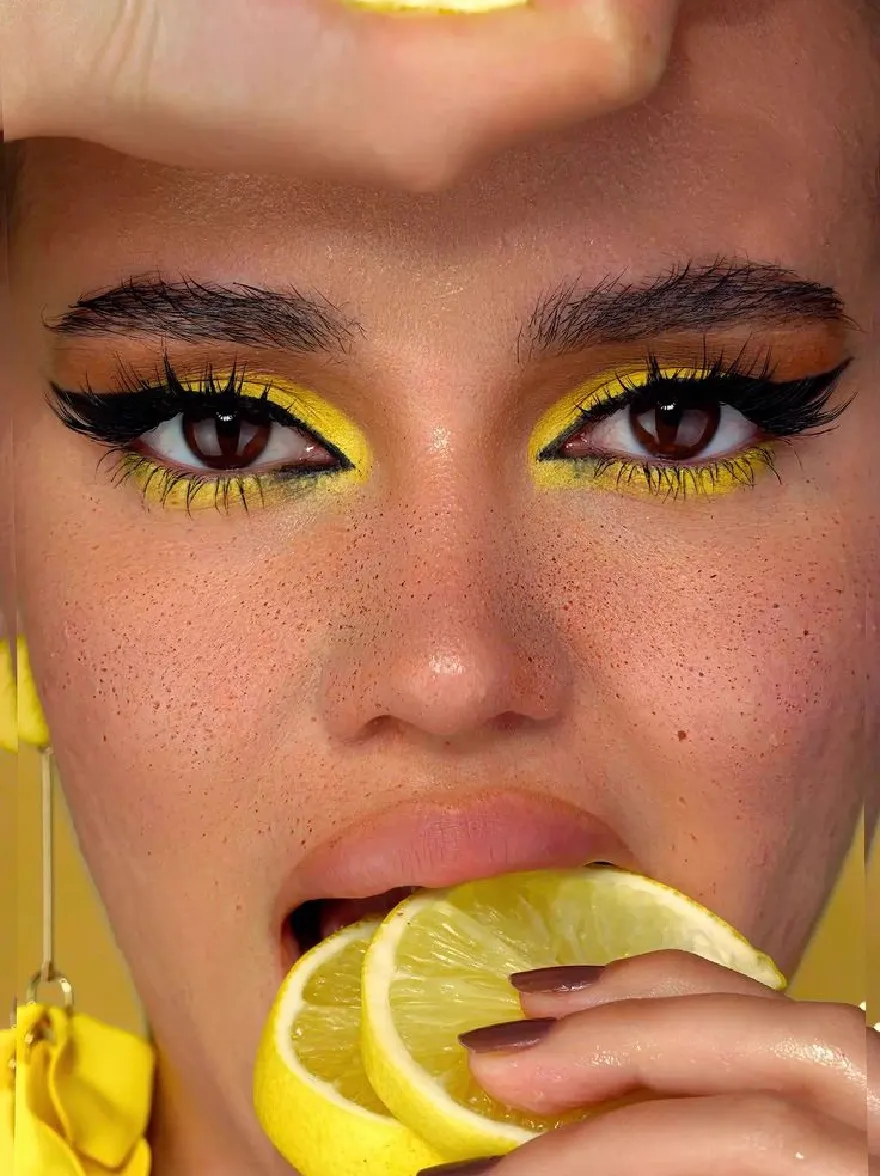 Русский промпт: Photo of a young woman with fair skin and freckles, holding two slices of lemon in front of her face. She has bright yellow eyeshadow, dark eyeliner, and dark nail polish. She is wearing large yellow earrings and has a playful expression. The background is a solid yellow color. The lighting is soft and natural, highlighting her features. The image has a high-quality, professional look. | Original prompt: Photo of a young woman with fair skin and freckles, holding two slices of lemon in front of her face. She has bright yellow eyeshadow, dark eyeliner, and dark nail polish. She is wearing large yellow earrings and has a playful expression. The background is a solid yellow color. The lighting is soft and natural, highlighting her features. The image has a high-quality, professional look.