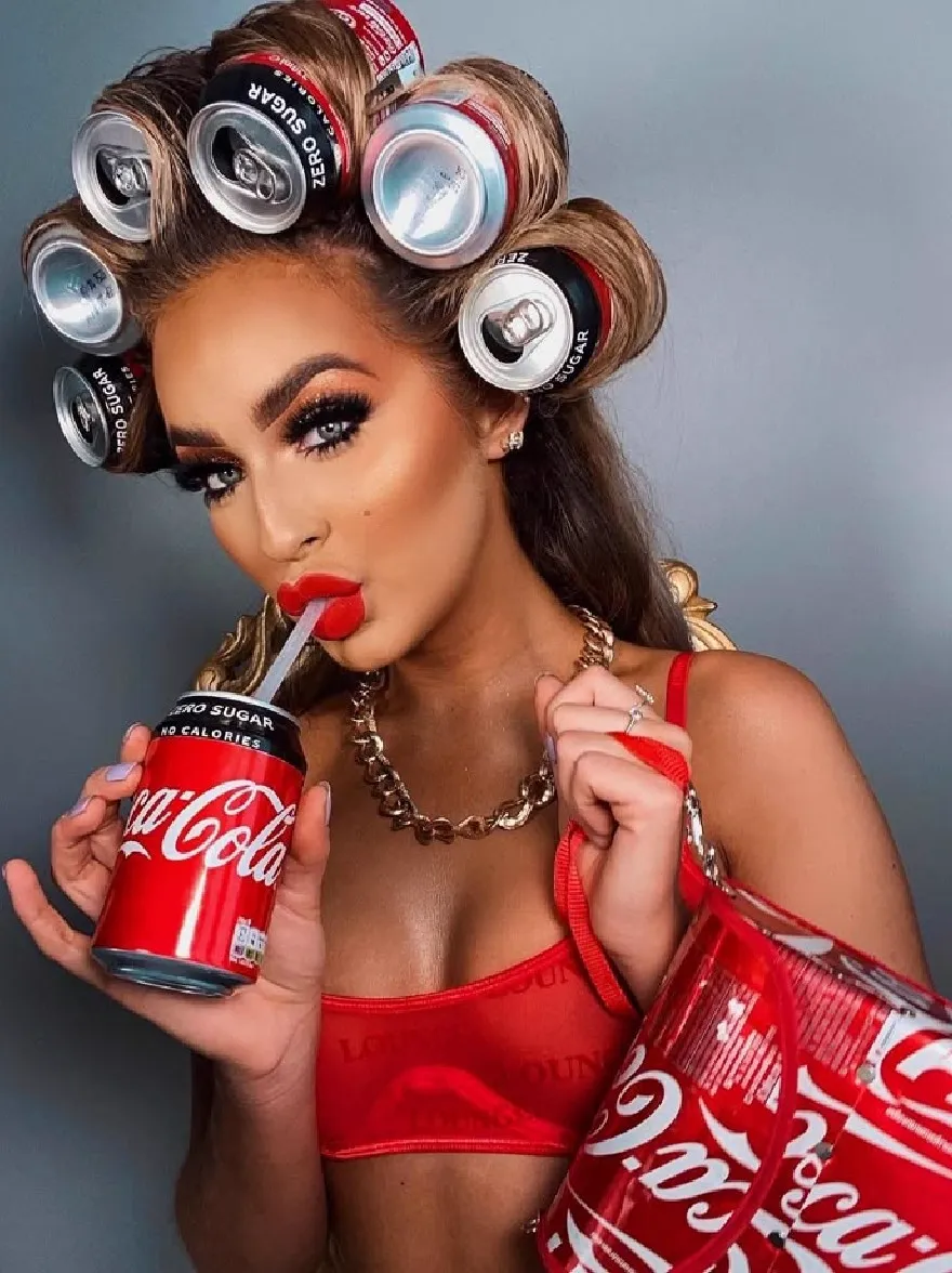 Русский промпт: Photo of a woman with dark skin, wearing a red crop top and holding a red Coca-Cola can in one hand and a red tote bag in the other. She has long, wavy brown hair styled in curlers with silver caps, and is wearing a gold chain necklace. Her makeup is bold and dramatic, with dark eyeliner and red lipstick. She is looking directly at the camera with a sultry expression. The background is a plain grey wall. | Original prompt: Photo of a woman with dark skin, wearing a red crop top and holding a red Coca-Cola can in one hand and a red tote bag in the other. She has long, wavy brown hair styled in curlers with silver caps, and is wearing a gold chain necklace. Her makeup is bold and dramatic, with dark eyeliner and red lipstick. She is looking directly at the camera with a sultry expression. The background is a plain grey wall.