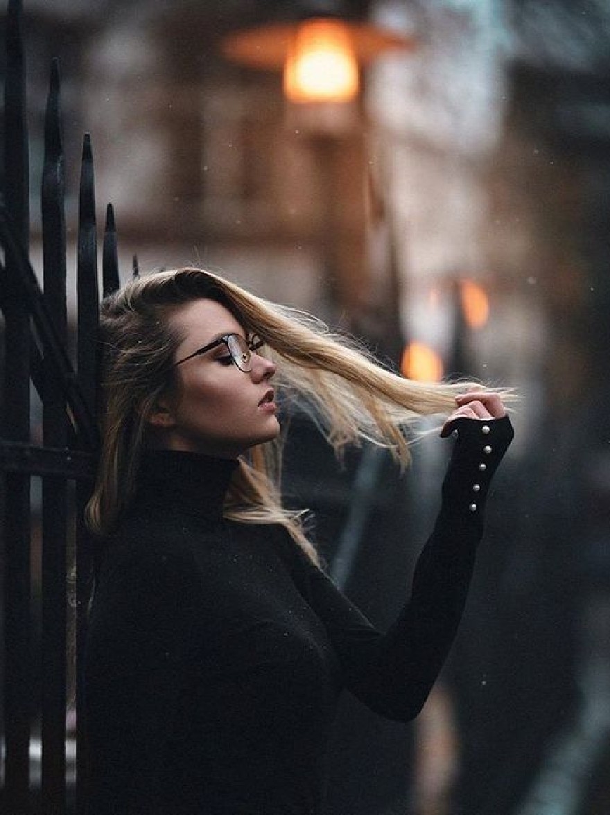 Русский промпт: Женщина с длинными светлыми волосами, стоящей в тускло освещенном переулке, одетый в черный свитер водолазки и круглые очки. Она отрегулирует свои волосы одной рукой, а другая рука опирается на забор. Фон размыт, с теплым оранжевым светом, освещающим сцену. Освещение мягкое и распространенное, создавая мирную атмосферу. У женщины серьезное выражение на лице. | Original prompt: A woman with long blonde hair standing in a dimly lit alleyway, wearing a black turtleneck sweater and round glasses. She is adjusting her hair with one hand, while the other hand is resting on the fence. The background is blurred, with a warm, orange light illuminating the scene. The lighting is soft and diffused, creating a peaceful atmosphere. The woman has a serious expression on her face.