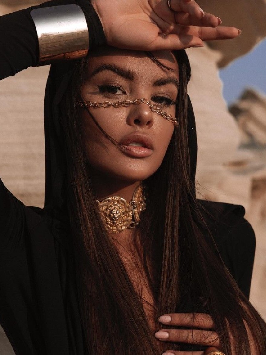 Русский промпт: Фотография красивой афро-американской женщины с длинными, прямыми темно-каштановыми волосами, в черном плащ с капюшоном и золотым кофером со сложными дизайнами. У нее серьезное выражение, и ее рука опирается на голову. Фон размыт, но, кажется, это наружная обстановка с ясным синим небом. Освещение теплое и естественное, подчеркивая ее черты. Изображение имеет высококачественное, профессиональное чувство. | Original prompt: Photo of a beautiful African-American woman with long, straight, dark brown hair, wearing a black hooded cloak and a gold choker with intricate designs. She has a serious expression, with her hand resting on her head. The background is blurred, but it appears to be an outdoor setting with a clear blue sky. The lighting is warm and natural, highlighting her features. The image has a high-quality, professional feel.