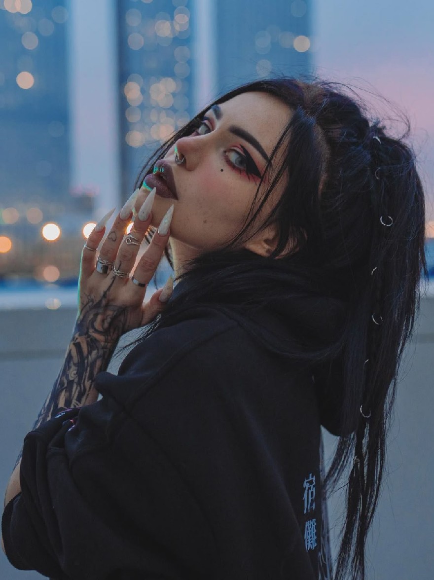 Русский промпт: Фотография молодой женщины с бледной кожей и длинными черными волосами, одетая в черную толстовку с белым логотипом на левом рукаве. У нее есть татуировка на левой руке и пирсинг на лице и шее. Она смотрит в сторону с серьезным выражением. Фон размыт, но это кажется городским пейзажем ночью с огнями. Освещение мягкое и теплое, создавая капризную атмосферу. Женщина расположена в центре рамы, с городским пейзажем на заднем плане. | Original prompt: Photo of a young woman with pale skin and long black hair, wearing a black hoodie with a white logo on the left sleeve. She has a tattoo on her left arm and piercings on her face and neck. She is looking to the side with a serious expression. The background is blurred, but it appears to be a cityscape at night with lights. The lighting is soft and warm, creating a moody atmosphere. The woman is positioned in the center of the frame, with the cityscape in the background.