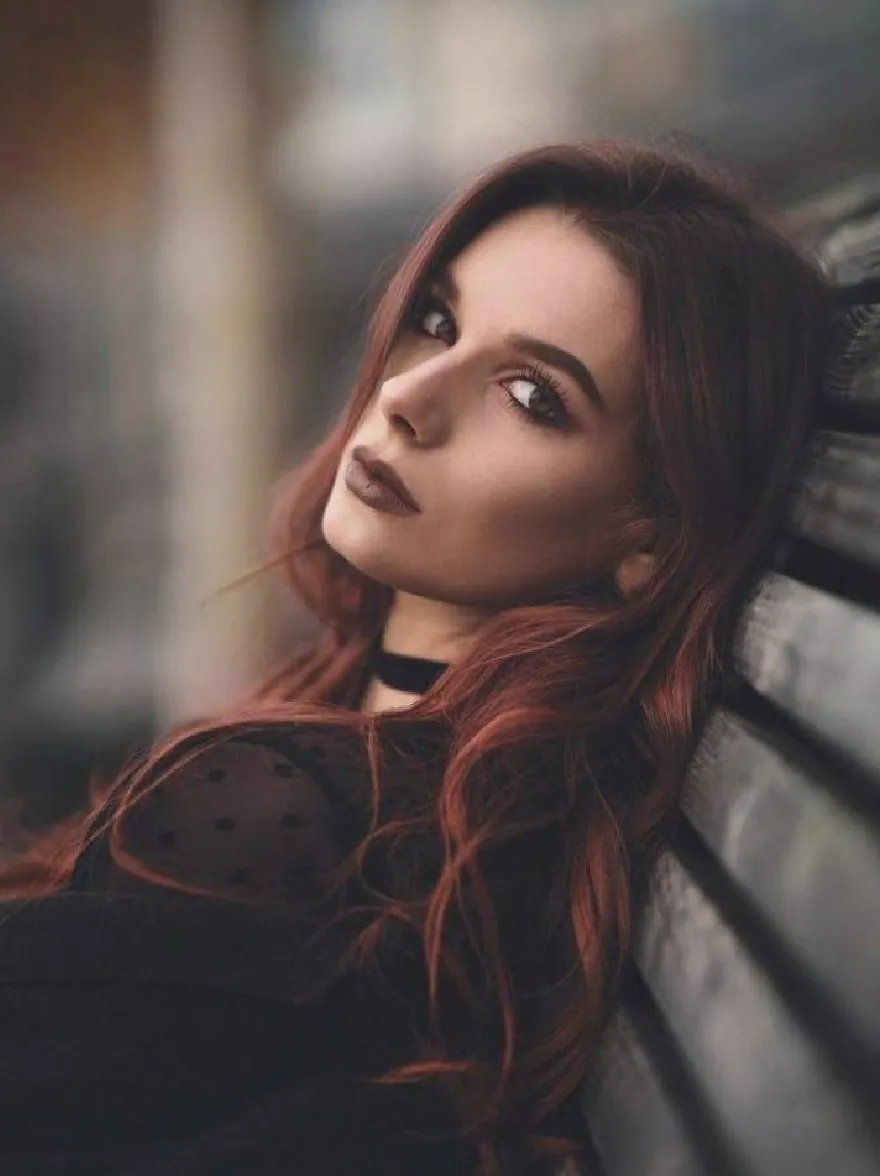 Русский промпт: Фото красивой женщины с длинными волнистыми рыжими волосами, отдыхая на деревянной скамейке. У нее есть честная кожа, темные глаза и тонкий дымный макияж глаз. На ней черный хокер и черный топ с горошками. Фон размыт, с деревенским, промышленным ощущением. Освещение мягкое и естественное, создавая безмятежную атмосферу. Женщина смотрит прямо на камеру с нейтральным выражением. Общее настроение мирное и созерцательное. | Original prompt: Photo of a beautiful woman with long, wavy red hair, resting head on a wooden bench. She has fair skin, dark eyes, and a subtle smoky eye makeup. She is wearing a black choker and a black top with polka dots. The background is blurred, with a rustic, industrial feel. The lighting is soft and natural, creating a serene atmosphere. The woman is looking directly at the camera with a neutral expression. The overall mood is peaceful and contemplative.