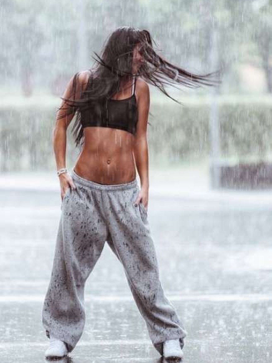 Русский промпт: Фотография молодой женщины с длинными, текущими черными волосами, в черном спортивном бюстгальтере и серых спортивных штанах, стоящей под дождем с руками на бедрах, глядя на землю. У нее приподнятое телосложение и носит белые кроссовки. Фон размыт, но, кажется, это наружная обстановка с деревьями и фонтаном. Освещение драматичное, с дождями падают вокруг нее. Изображение высококачественное и имеет водяной знак в правом нижнем углу. | Original prompt: Photo of a young woman with long, flowing black hair, wearing a black sports bra and grey sweatpants, standing in the rain with her hands on her hips, looking down at the ground. She has a fit physique and is wearing white sneakers. The background is blurred, but it appears to be an outdoor setting with trees and a fountain. The lighting is dramatic, with raindrops falling around her. The image is high quality and has a watermark in the bottom right corner.