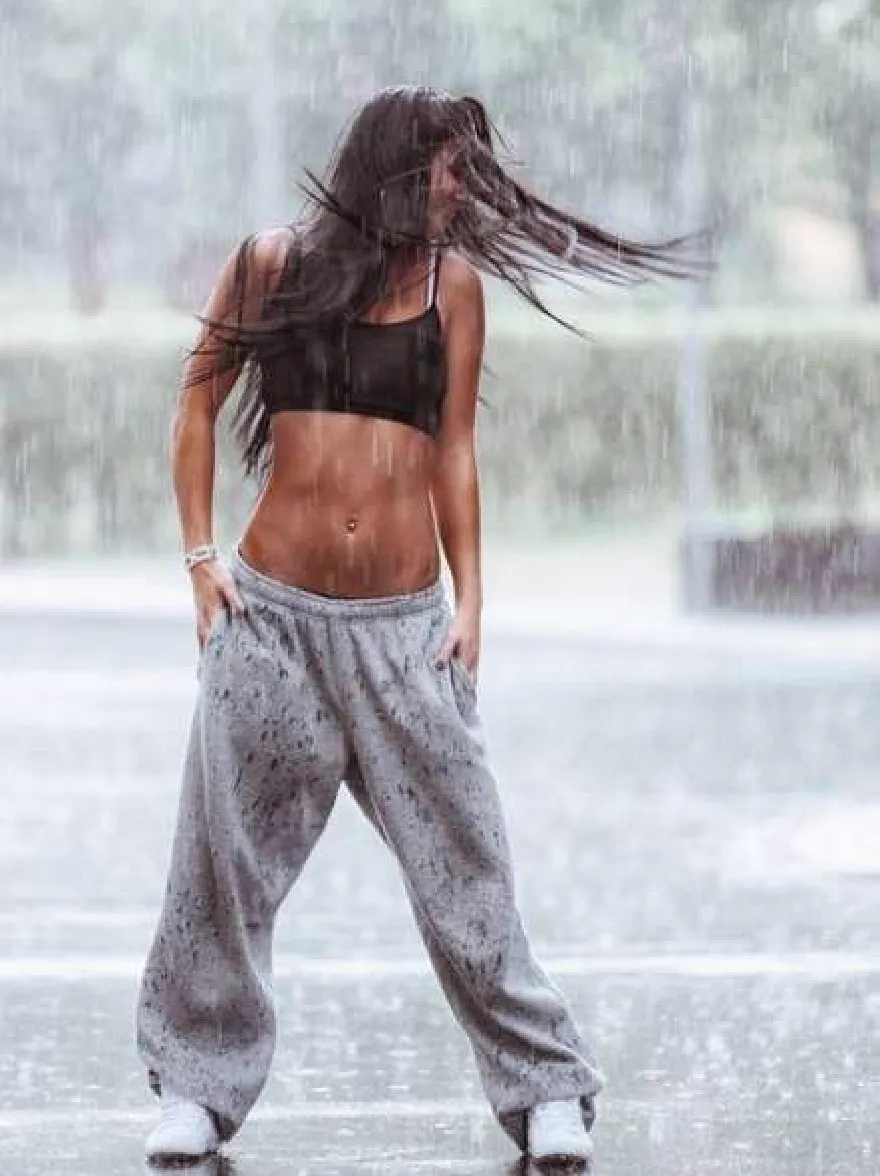 Русский промпт: Фотография молодой женщины с длинными, текущими черными волосами, в черном спортивном бюстгальтере и серых спортивных штанах, стоящей под дождем с руками на бедрах, глядя на землю. У нее приподнятое телосложение и носит белые кроссовки. Фон размыт, но, кажется, это наружная обстановка с деревьями и фонтаном. Освещение драматичное, с дождями падают вокруг нее. Изображение высококачественное и имеет водяной знак в правом нижнем углу. | Original prompt: Photo of a young woman with long, flowing black hair, wearing a black sports bra and grey sweatpants, standing in the rain with her hands on her hips, looking down at the ground. She has a fit physique and is wearing white sneakers. The background is blurred, but it appears to be an outdoor setting with trees and a fountain. The lighting is dramatic, with raindrops falling around her. The image is high quality and has a watermark in the bottom right corner.
