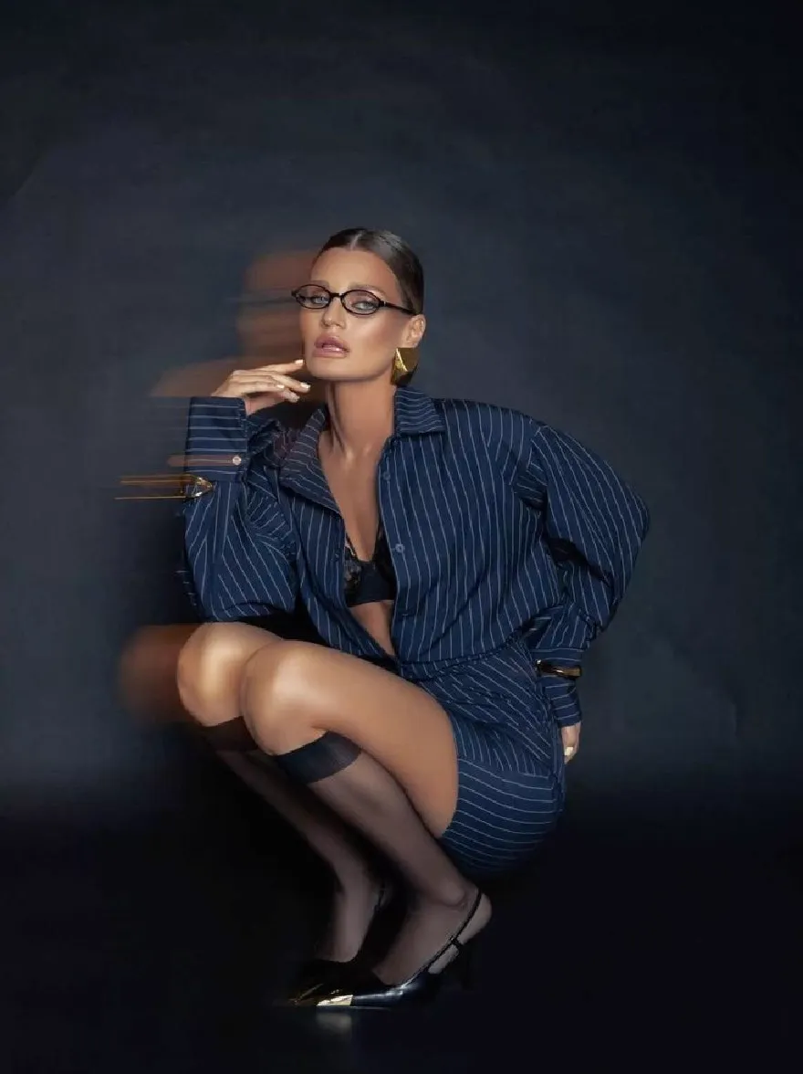 Русский промпт: Photo of a woman with dark skin, wearing a navy blue pinstripe suit with gold accents, black thigh-high stockings, and black high heels. She has short, dark hair styled in a sleek, straight look. She is squatting down with her hands resting on her knees, looking directly at the camera with a serious expression. The background is a dark, textured wall. The lighting is dramatic, casting a soft glow on her face and body. The overall mood is sophisticated and edgy. | Original prompt: Photo of a woman with dark skin, wearing a navy blue pinstripe suit with gold accents, black thigh-high stockings, and black high heels. She has short, dark hair styled in a sleek, straight look. She is squatting down with her hands resting on her knees, looking directly at the camera with a serious expression. The background is a dark, textured wall. The lighting is dramatic, casting a soft glow on her face and body. The overall mood is sophisticated and edgy.