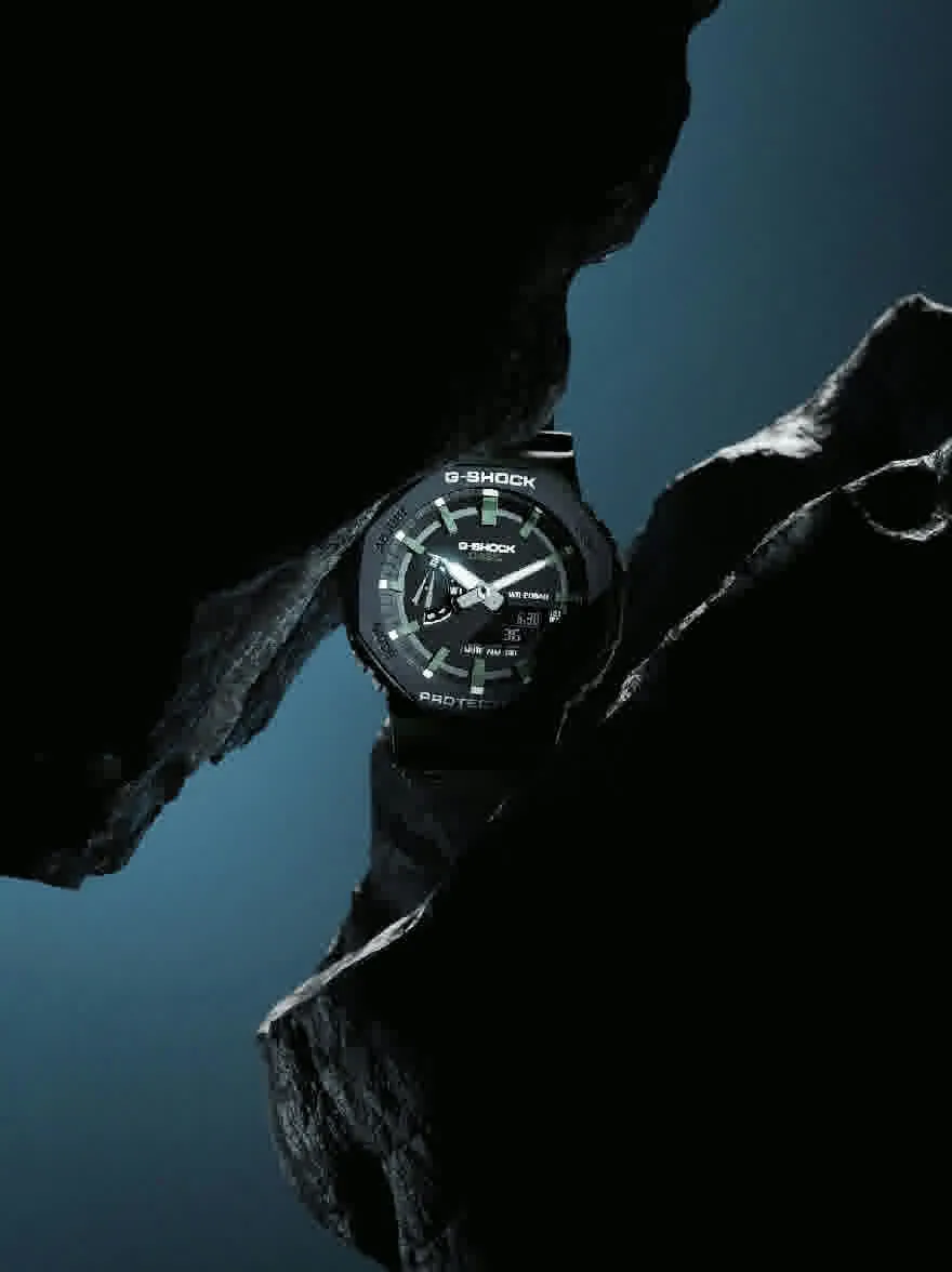 Русский промпт: Фотография сбоку Casio G-Shock G-Steel G-Squad, расположенная на темной скалистой скале с синим градиентным фоном. Часы имеют черное лицо со светящимися руками и маркерами, а также черный резиновый ремешок. Текст "G-Shock" заметно отображается в верхней части лица. Изображение снимается под низким углом, глядя на часы. Освещение драматичное, подчеркивая гладкий дизайн часов и текстуру камня. Общее настроение темное и загадочное. | Original prompt: A photograph of a Casio G-Shock G-Steel G-Squad watch from the side, set against a dark, rocky cliff with a blue gradient background. The watch has a black face with luminous hands and markers, and a black rubber strap. The text "G-Shock" is prominently displayed at the top of the watch face. The image is taken from a low angle, looking up at the watch. The lighting is dramatic, highlighting the watch's sleek design and the texture of the rock. The overall mood is dark and mysterious.